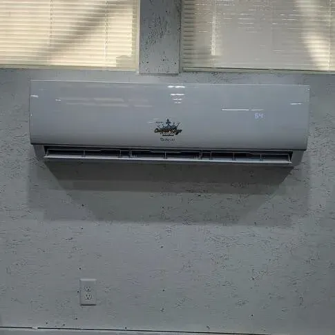 White air conditioner mounted on a gray concrete wall, below a window with blinds.