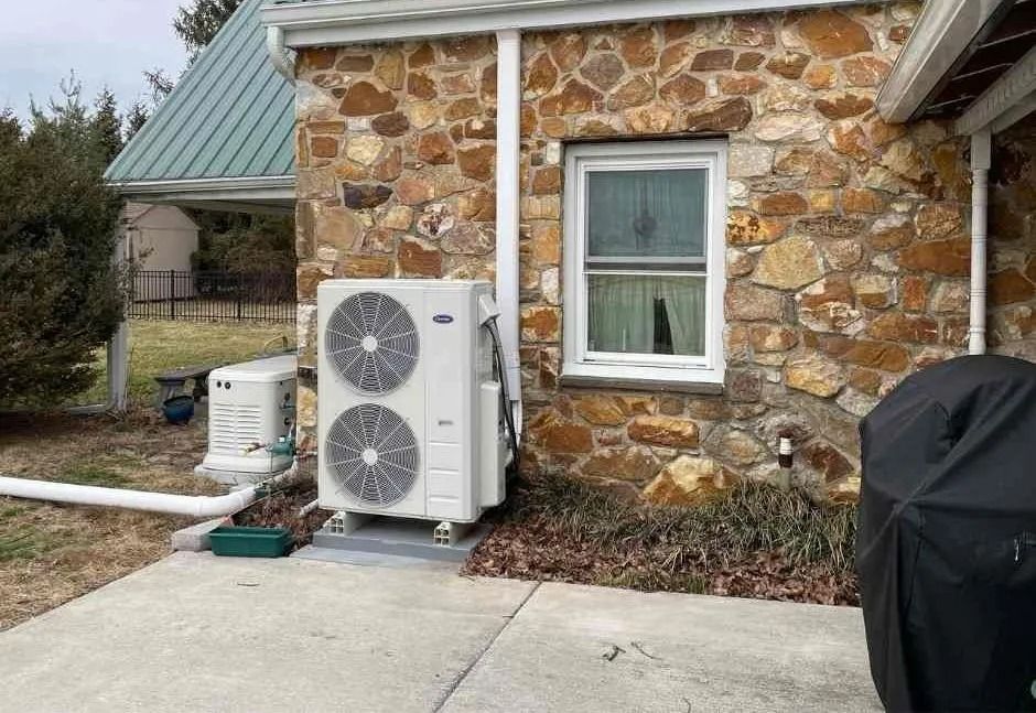 Air conditioning unit mounted on blocks against a brick building, snowy ground.