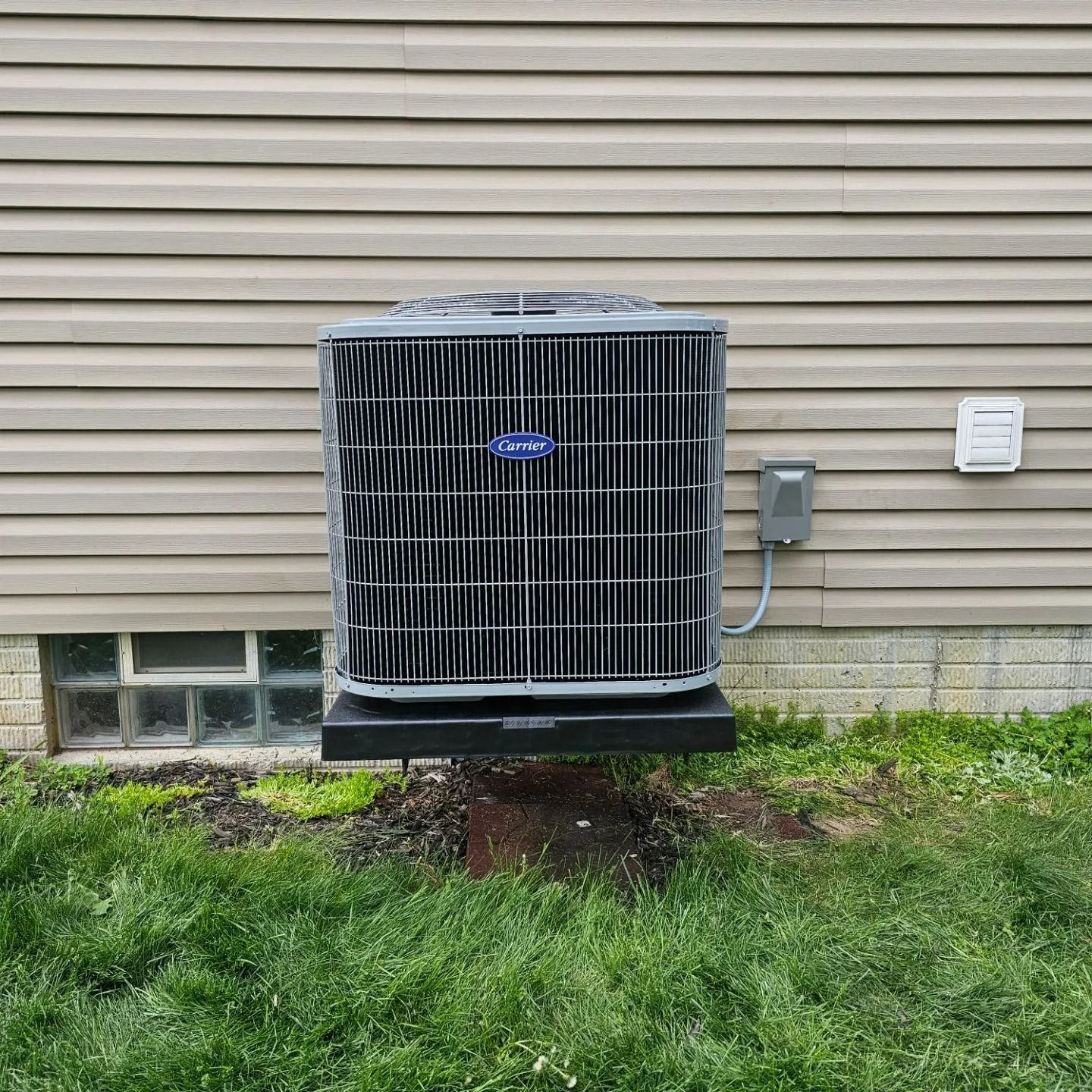 Carrier air conditioning unit mounted on a black base against a tan house wall, with green grass in foreground.