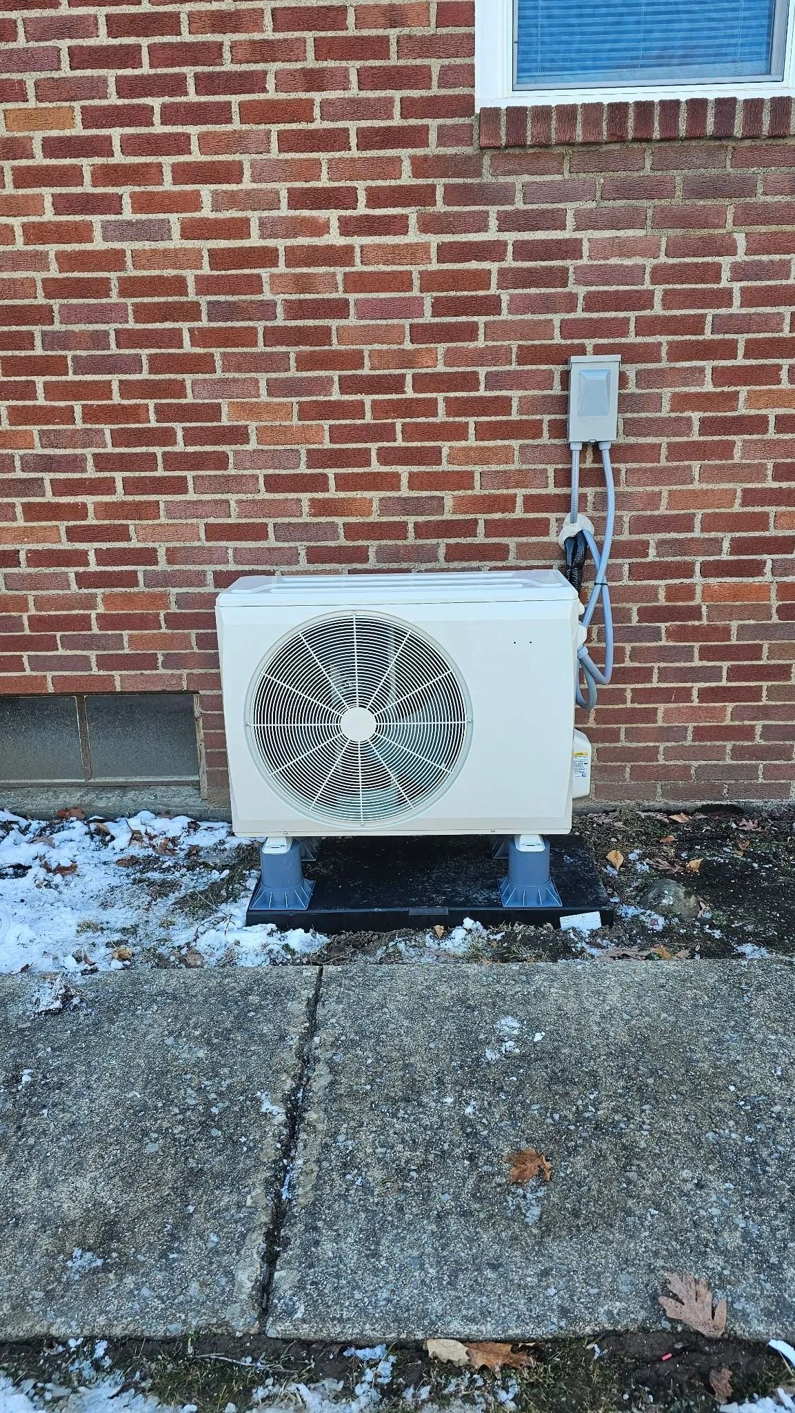Outdoor AC unit against a brick wall, resting on black platform; snow on the ground.