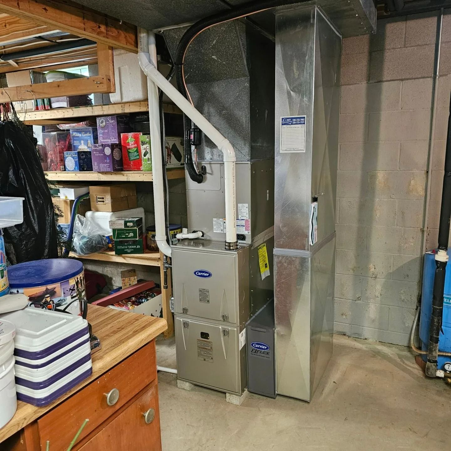 Furnace unit in a basement, with ductwork and nearby shelving with items.
