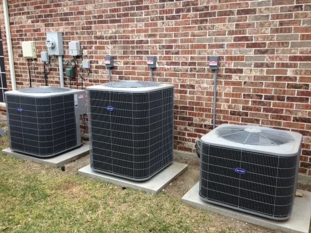 Two outdoor air conditioning units on a bed of gravel near a brown wooden fence.