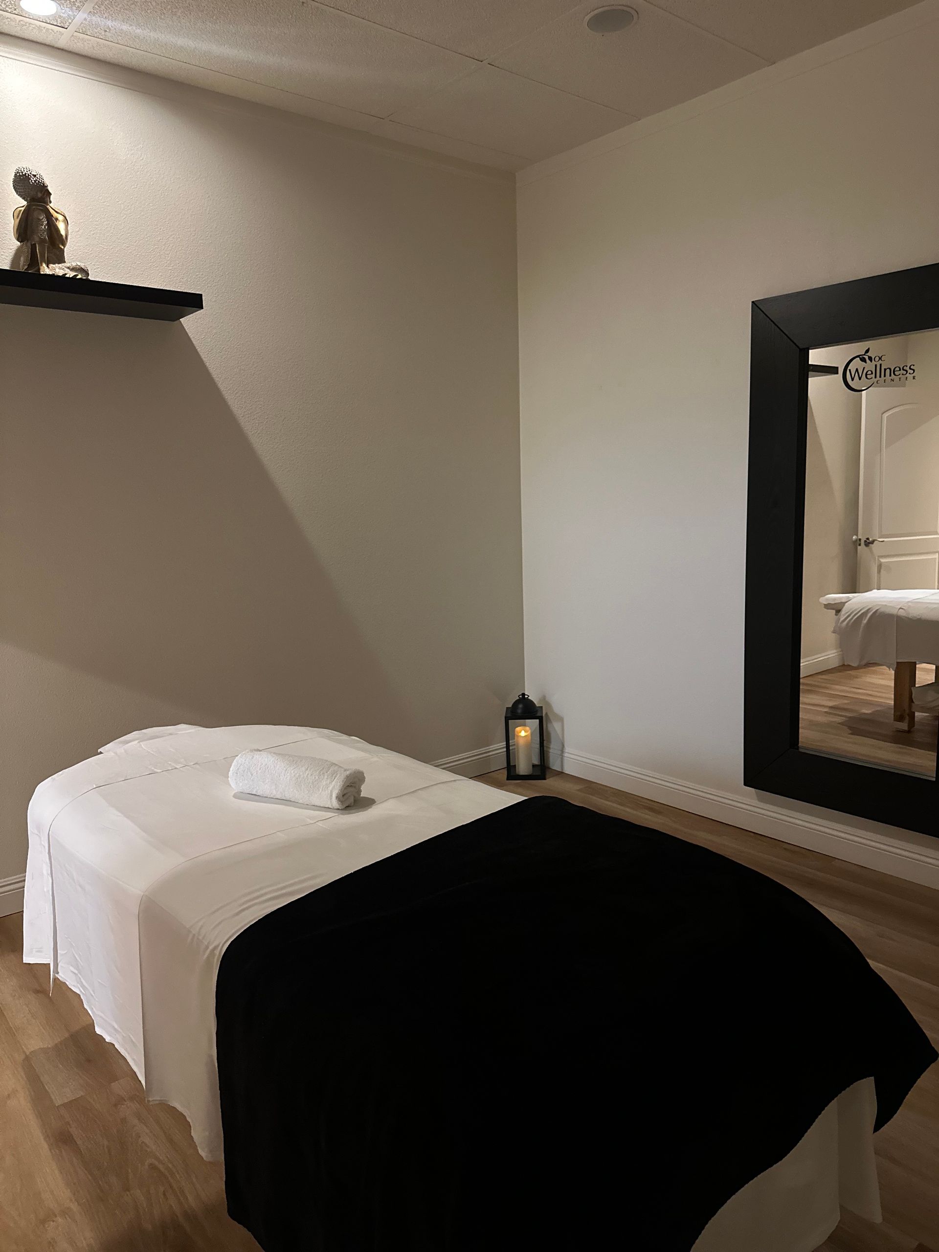 Massage room: Massage table with white linens and black blanket, a large mirror, and a small shelf with decor.