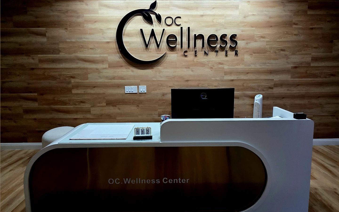 Reception desk in OC Wellness Clinic. Wooden wall with logo above, desk is white and gold.
