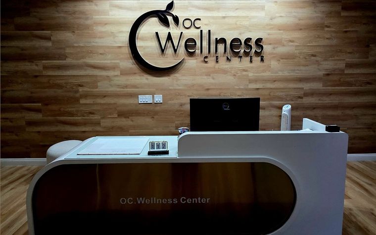 Reception desk in OC Wellness Clinic. Wooden wall with logo above, desk is white and gold.