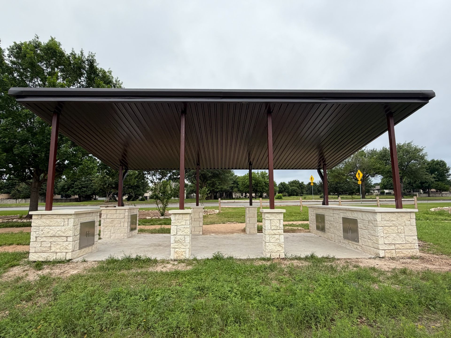Open-air pavilion with a dark metal roof and stone pillars in a grassy park area.