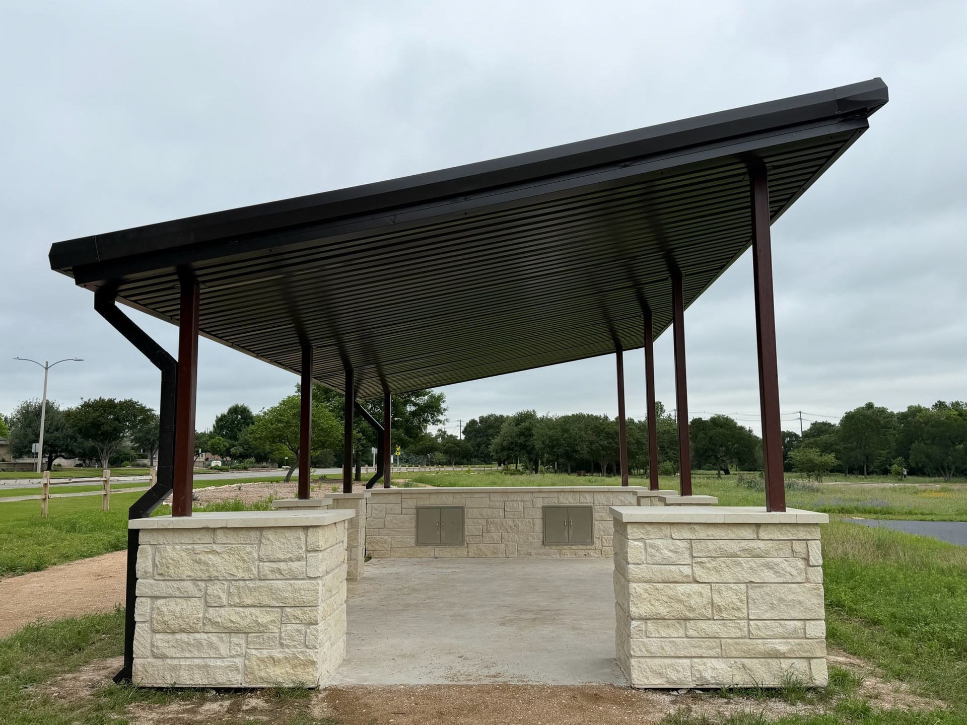 Open-air pavilion with a curved metal roof and stone benches in a grassy park