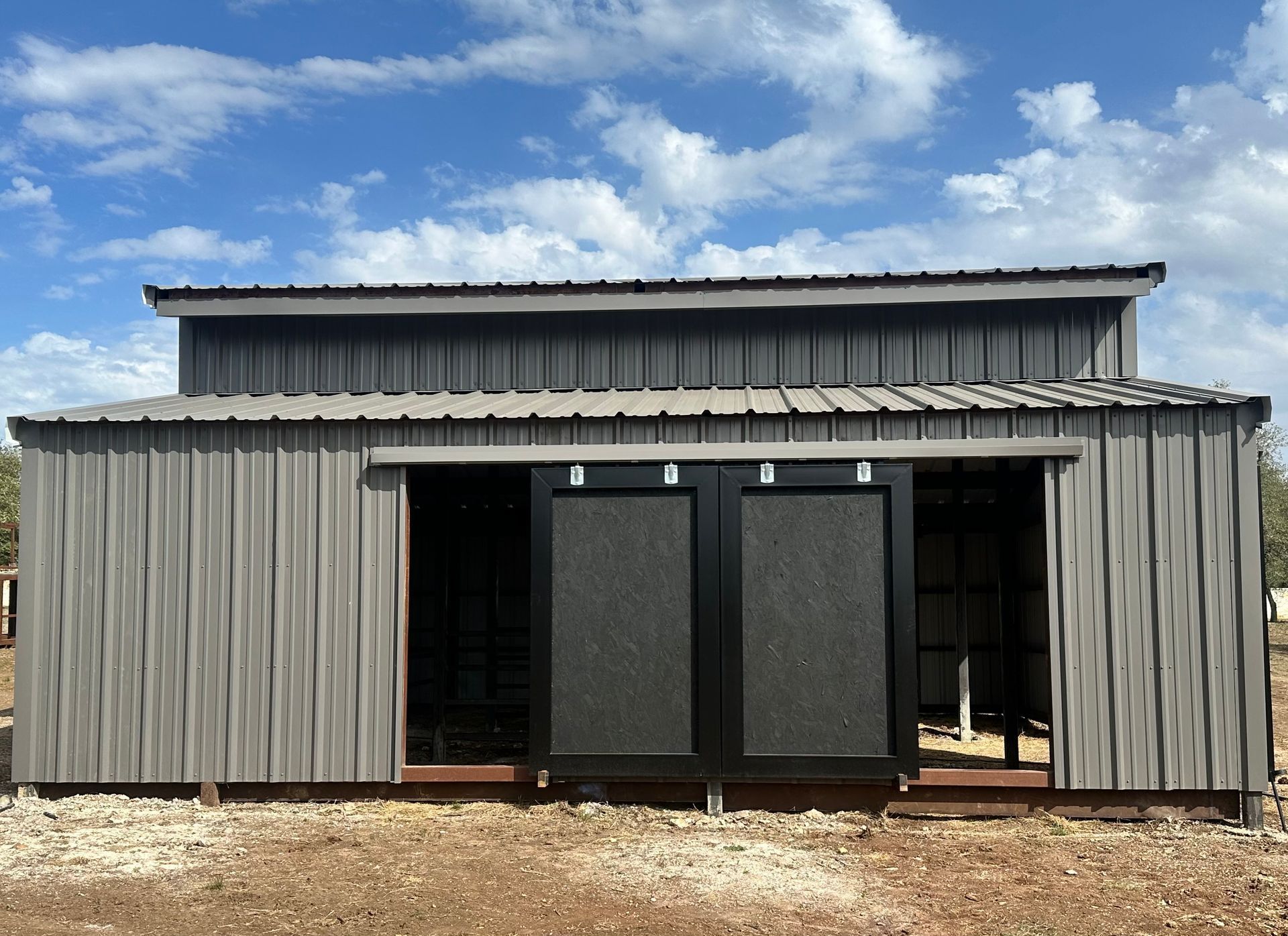 Gray metal barn with sliding black doors, set against a blue sky with clouds.