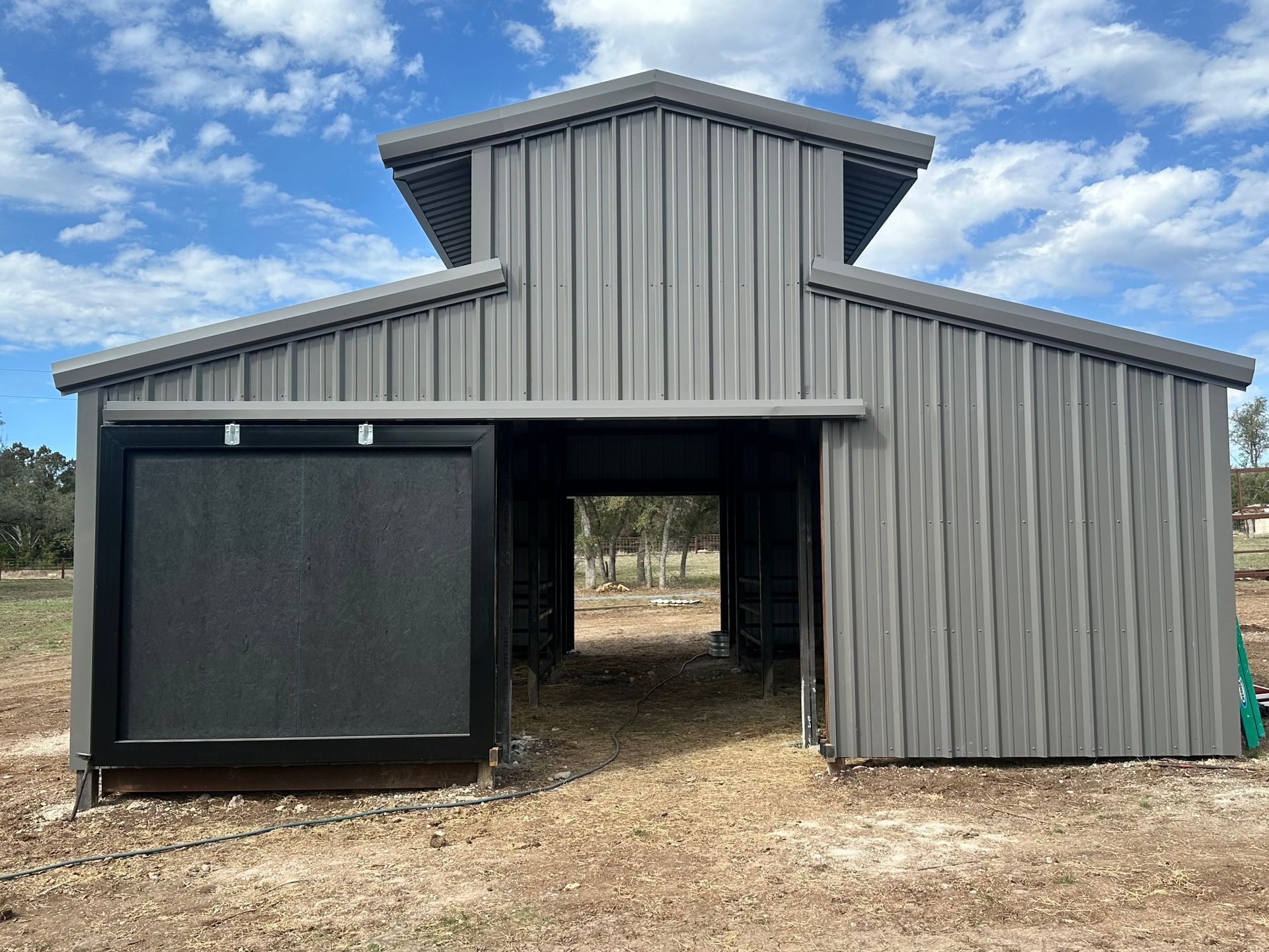 Gray metal barn with a sliding black door and open entryway against a cloudy sky.
