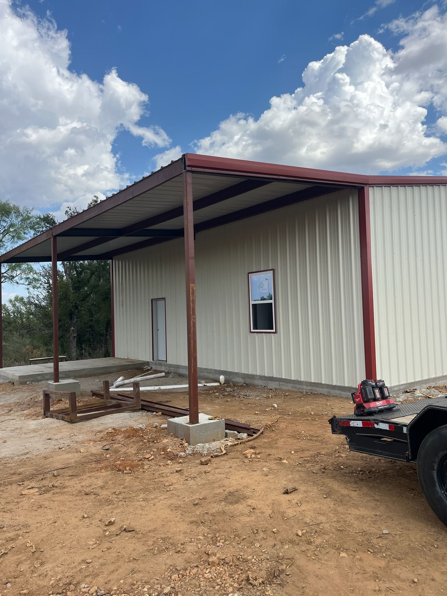 Small metal building with a red-roofed porch on dirt ground under a cloudy sky.