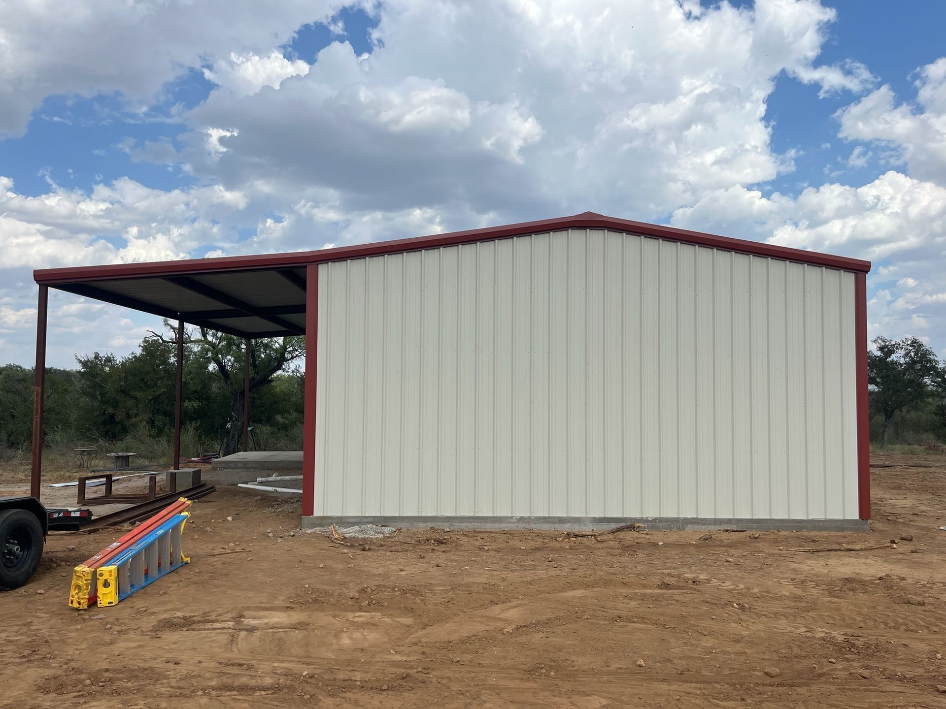 Large beige metal building with a red roof beside an open-sided shed on a dirt lot under a cloudy sky