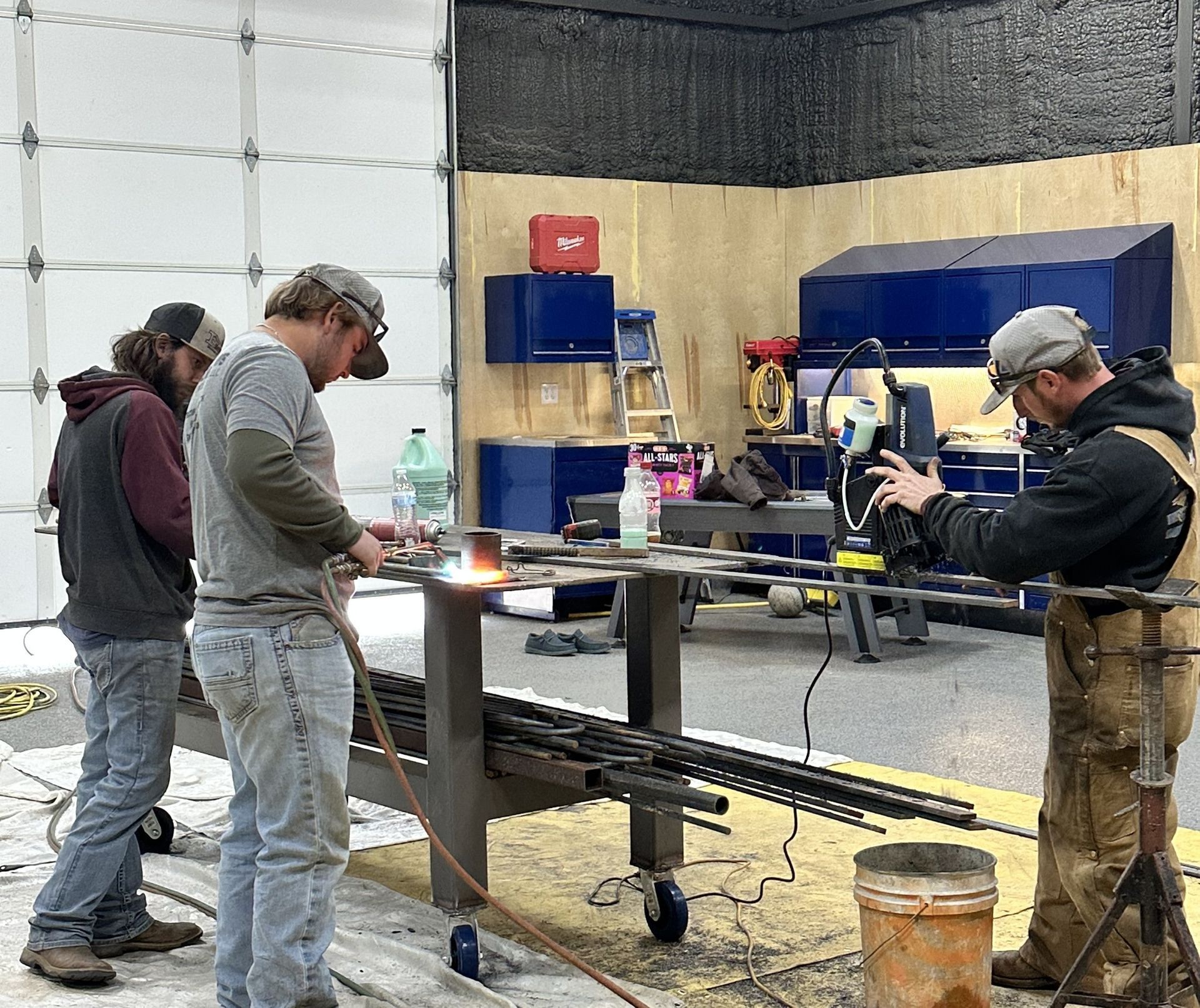 Three workers welding metal rods in a garage; one wears safety glasses, another films.