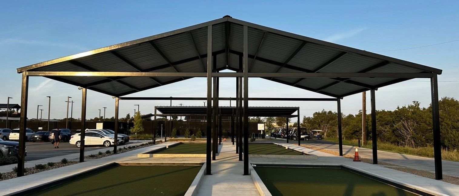 Metal-framed bocce ball courts with a covered roof in a park, with cars parked nearby on a clear day.