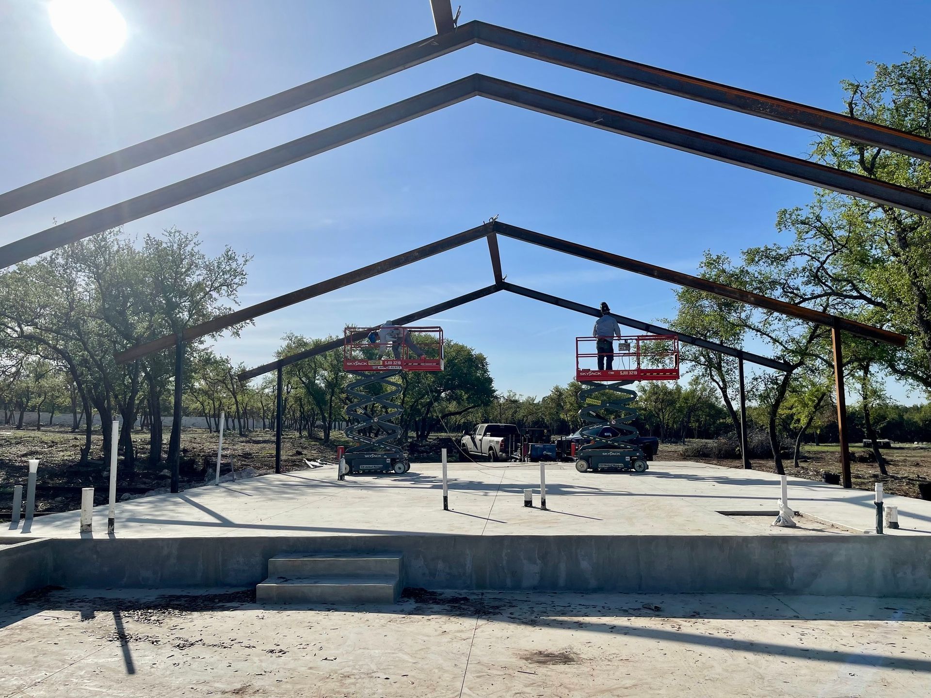 Steel building frame being erected on a concrete foundation; workers on lifts. Sunny day.