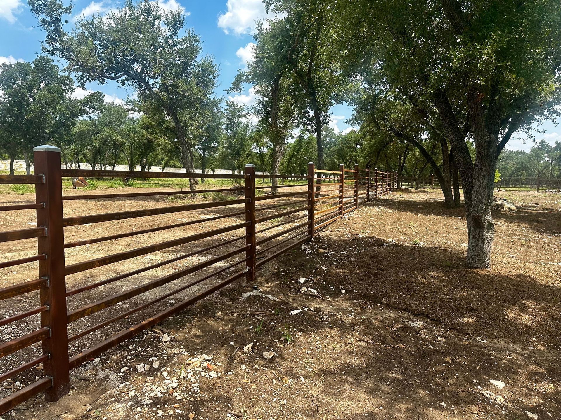 Metal fence in a rural setting, brown and tan colors, trees and blue sky.