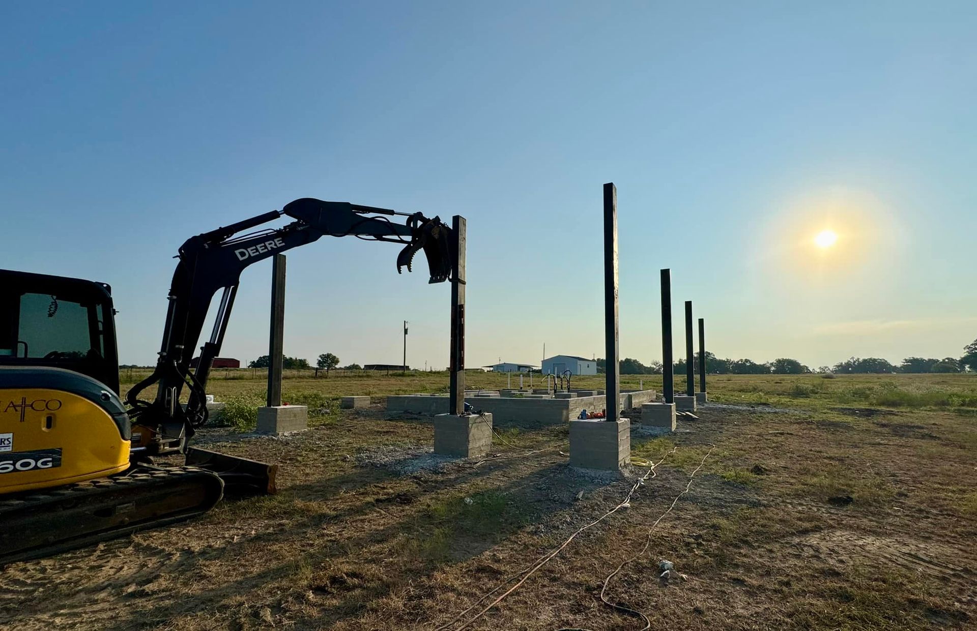 Yellow excavator placing a pole into a concrete base on a field. Sun shining in the background.