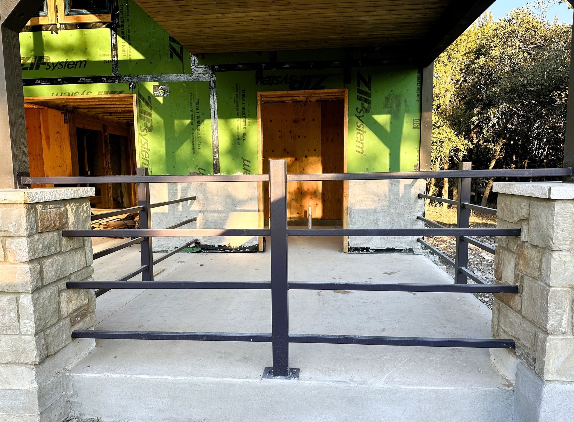 A porch with black metal railing, stone columns, and concrete steps. Construction underway.