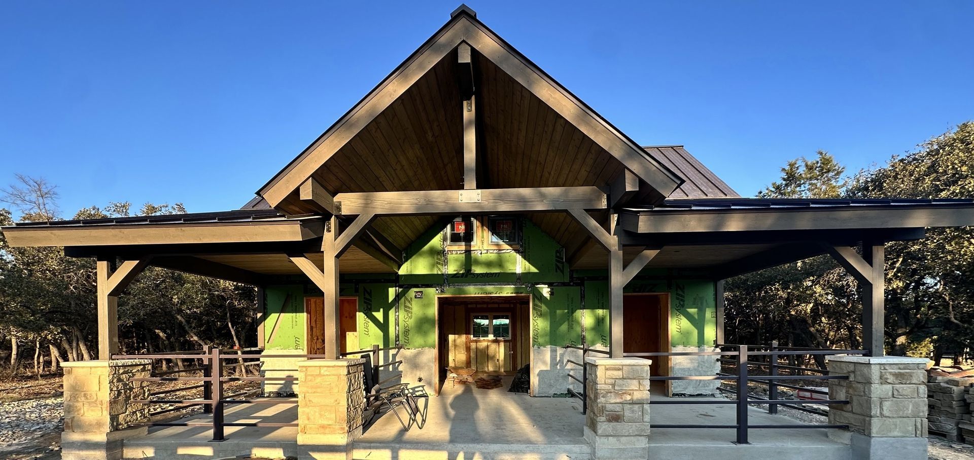 A wooden pavilion with a gabled roof stands in a park. Green walls and brown beams with stone columns.