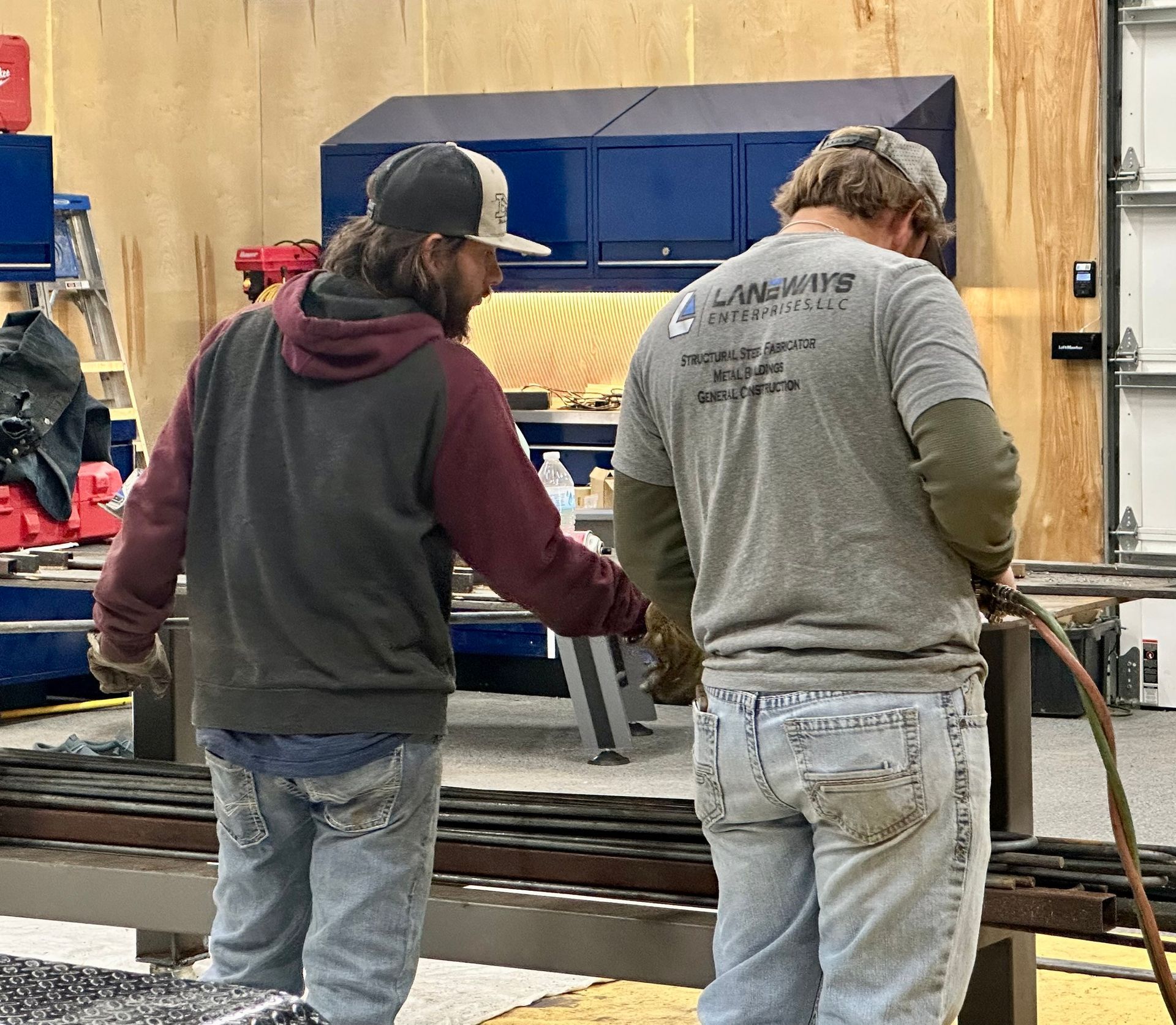 Two men in a workshop, one pointing while the other works with a torch on metal bars.