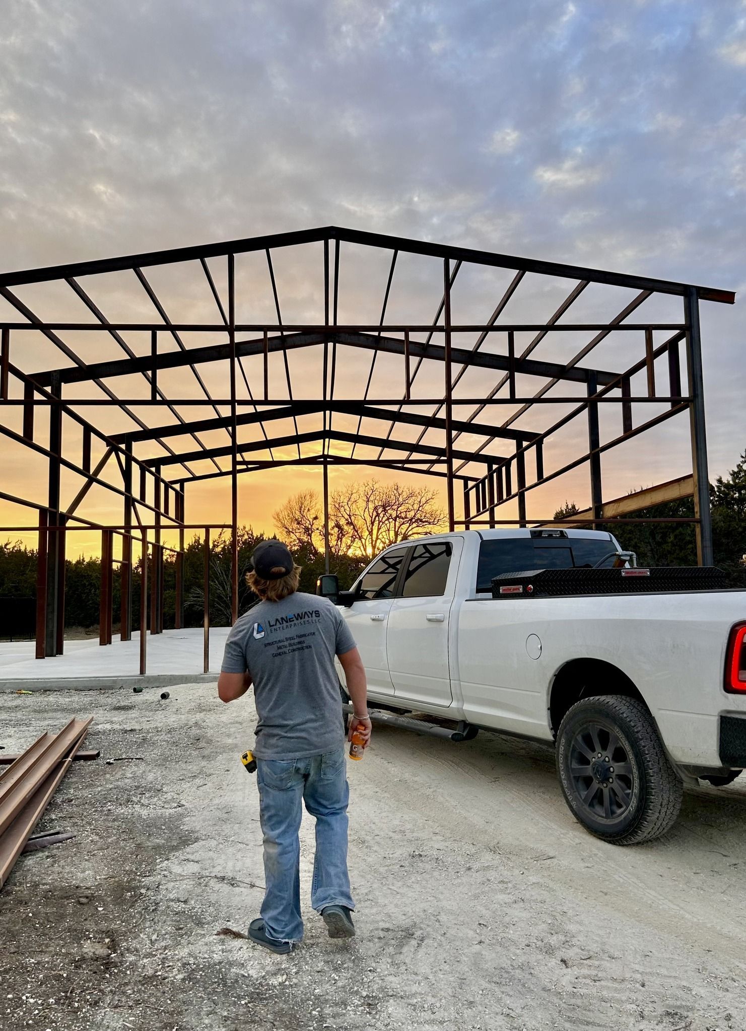 A man stands near a white truck, in front of a metal building frame, at dusk.