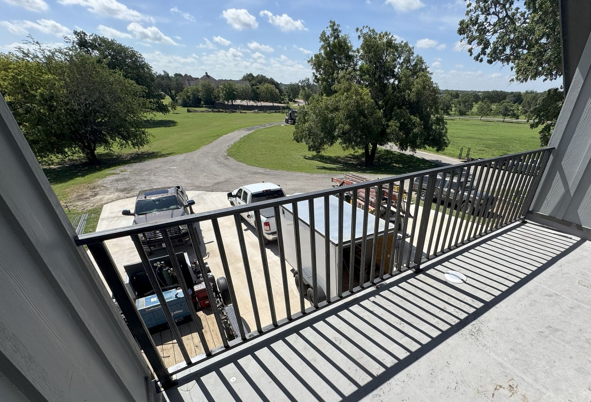 View from a balcony with black railings. Cars, a gravel road, and green fields are visible.