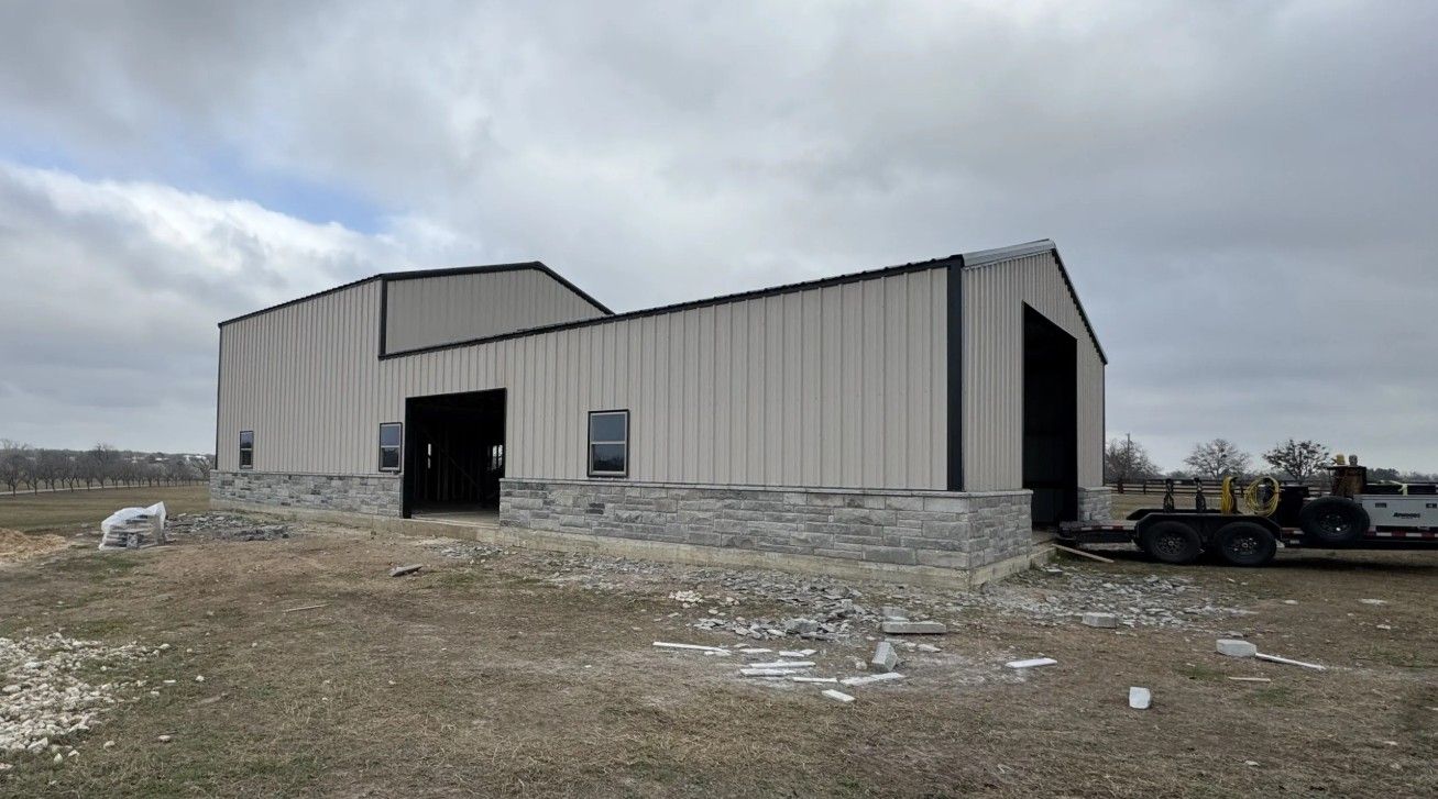 Barn with light-colored metal siding and a stone base, under an overcast sky. A trailer is parked nearby.