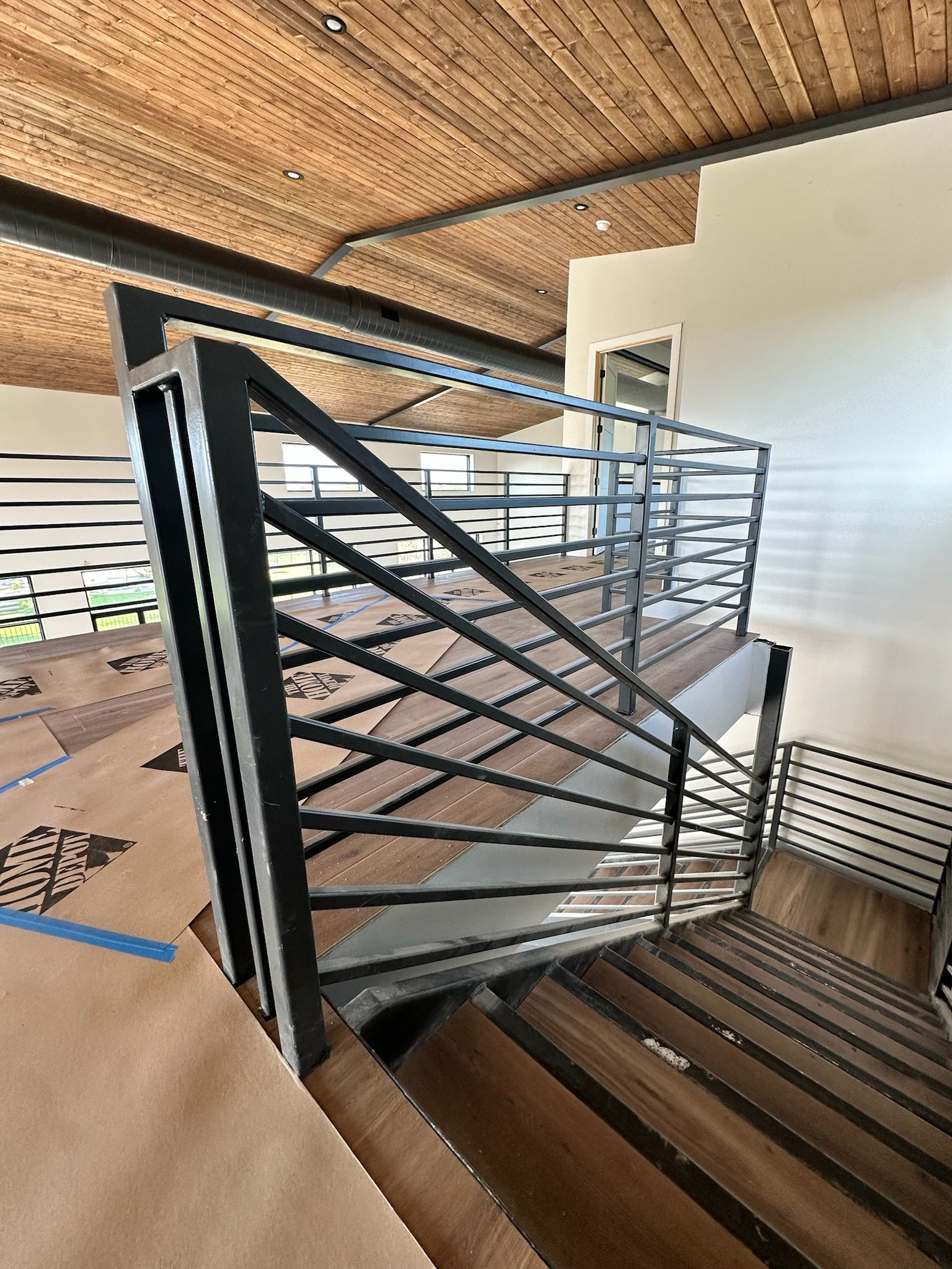 Staircase with modern black metal railing and wooden steps, leading up and down. Brown ceiling.