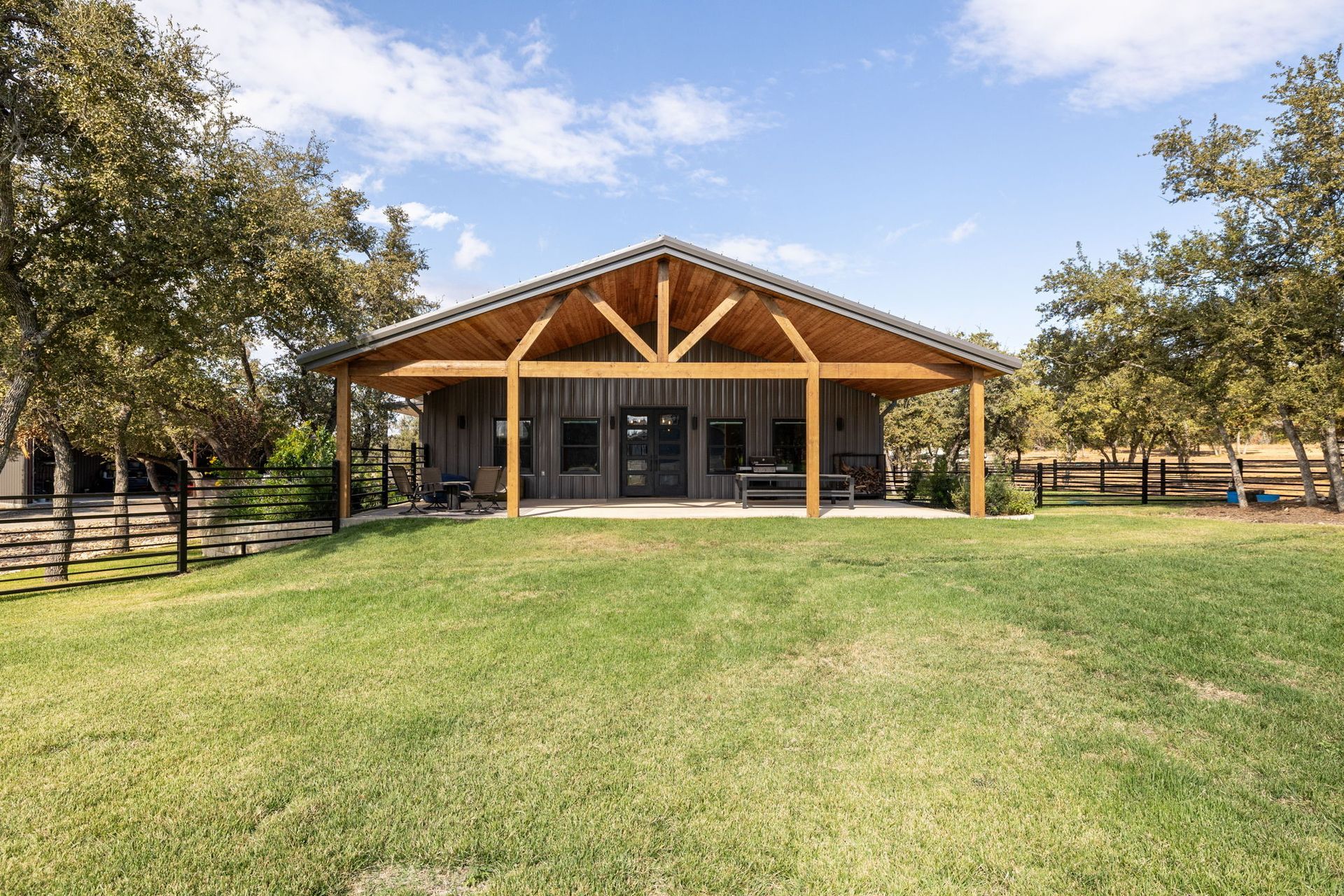 Barn-style building with a porch, green lawn, trees, and a fence under a blue sky.