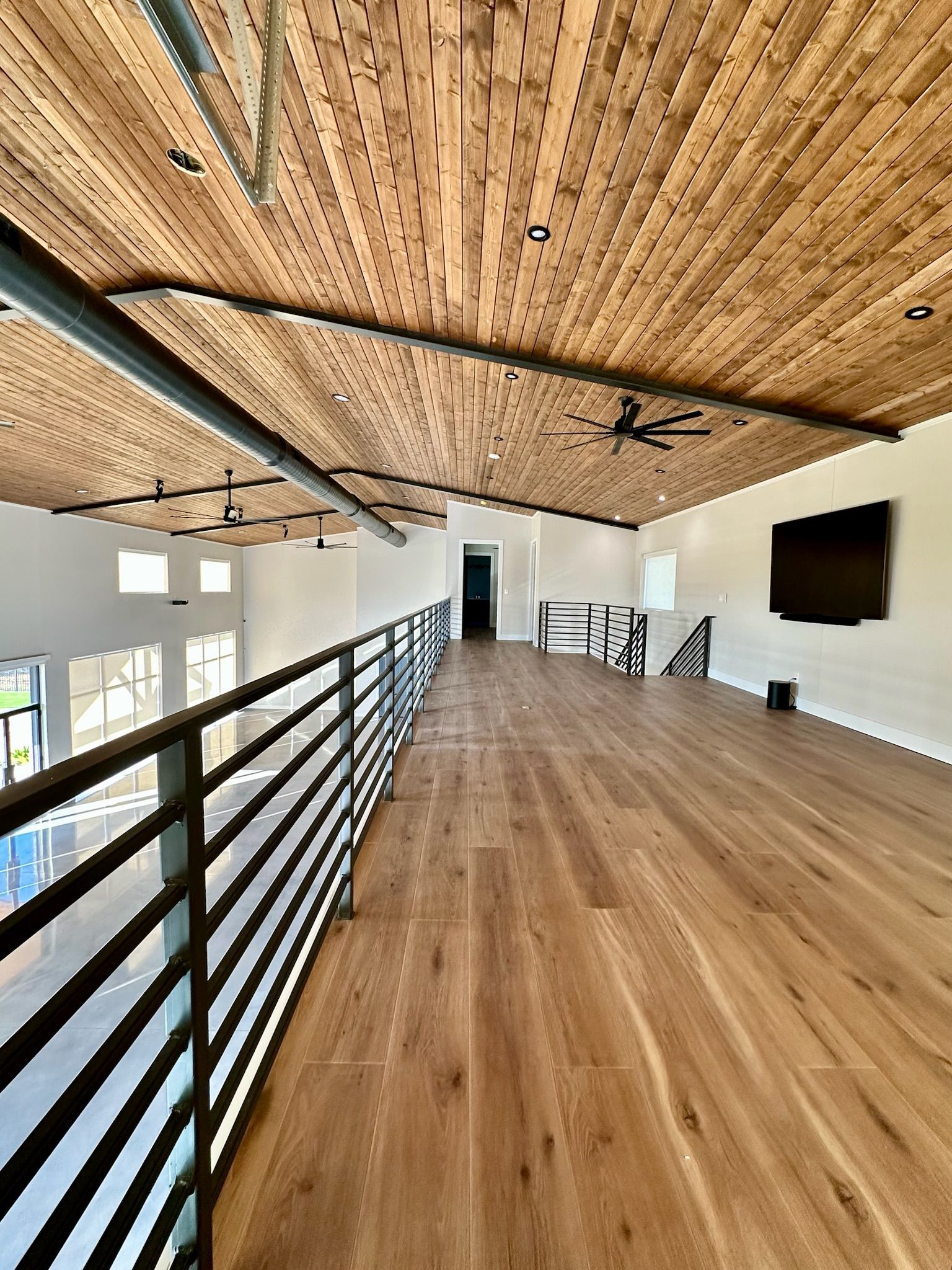 Interior view of a modern loft with wood ceiling and floors, black metal railing, and a large TV.
