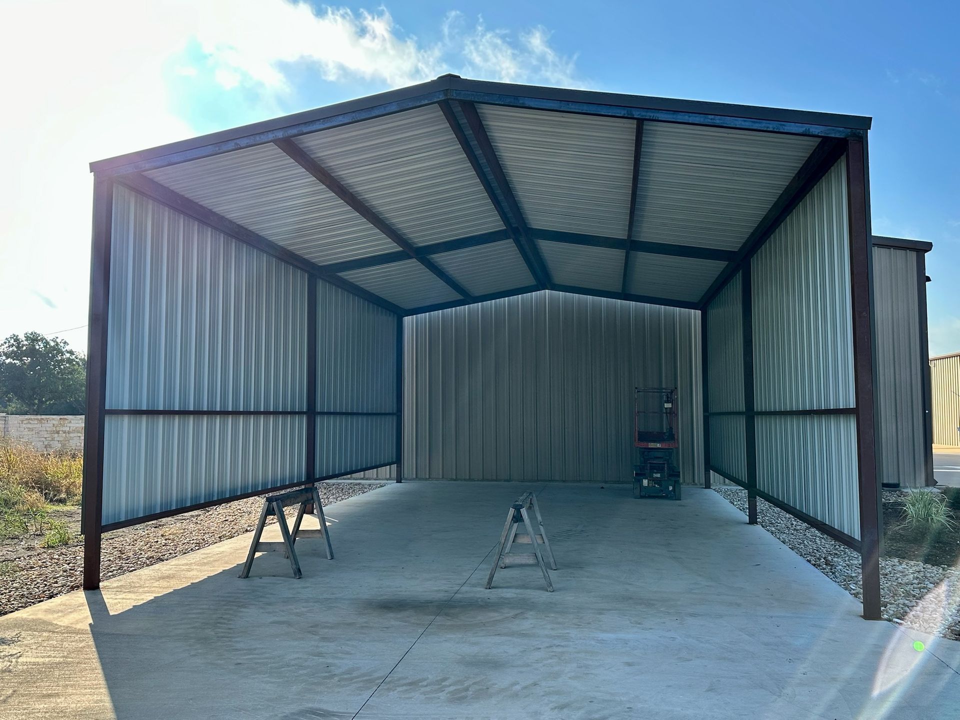 Metal carport with a concrete floor, two sawhorses, and blue sky.