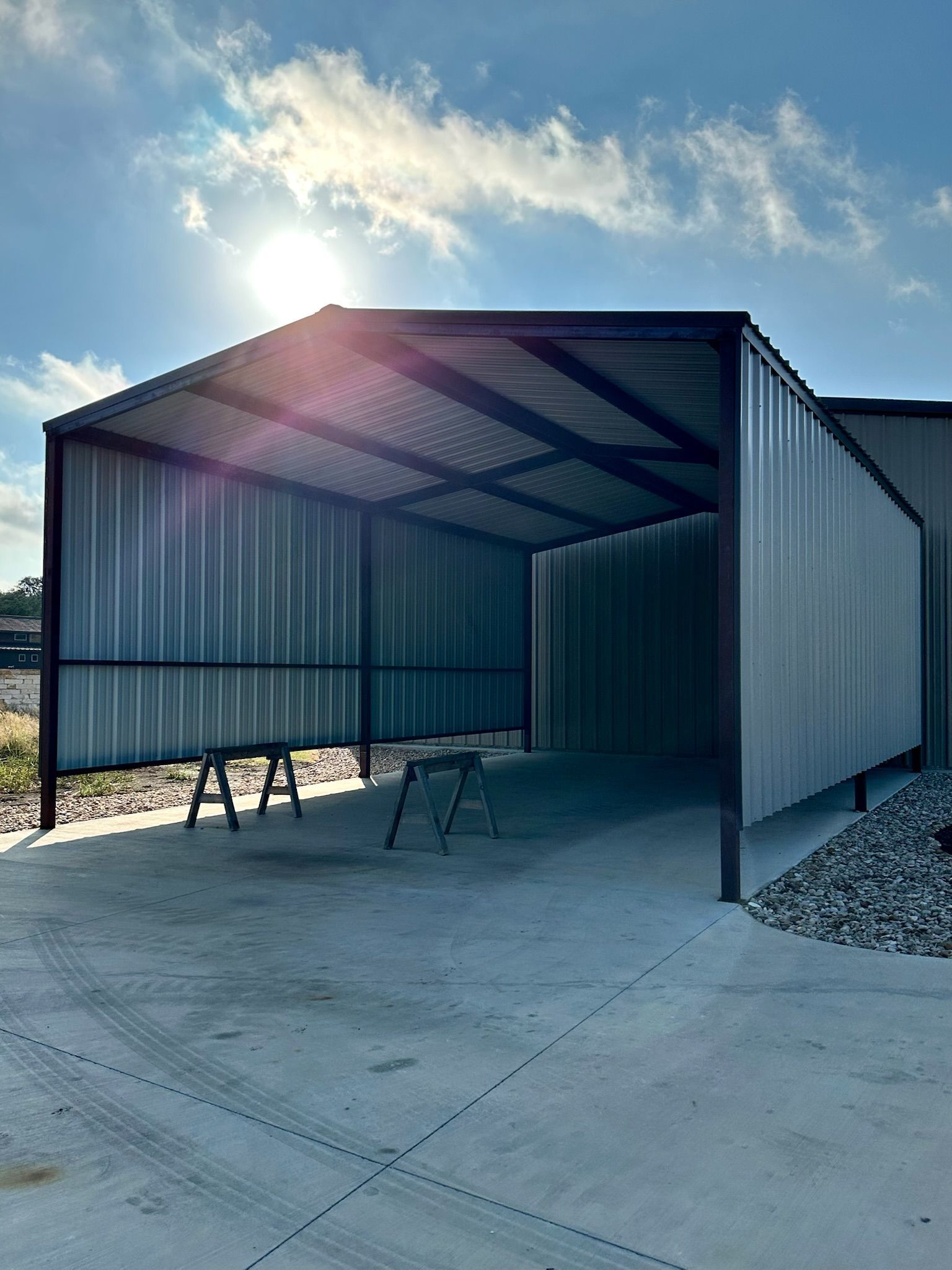 Metal carport with a concrete floor, two sawhorses, and sun shining.