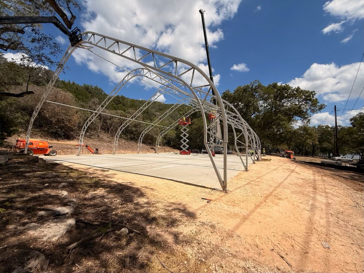 Steel pavilion frame under construction on a concrete pad beside a dirt road, with trees and blue sky.