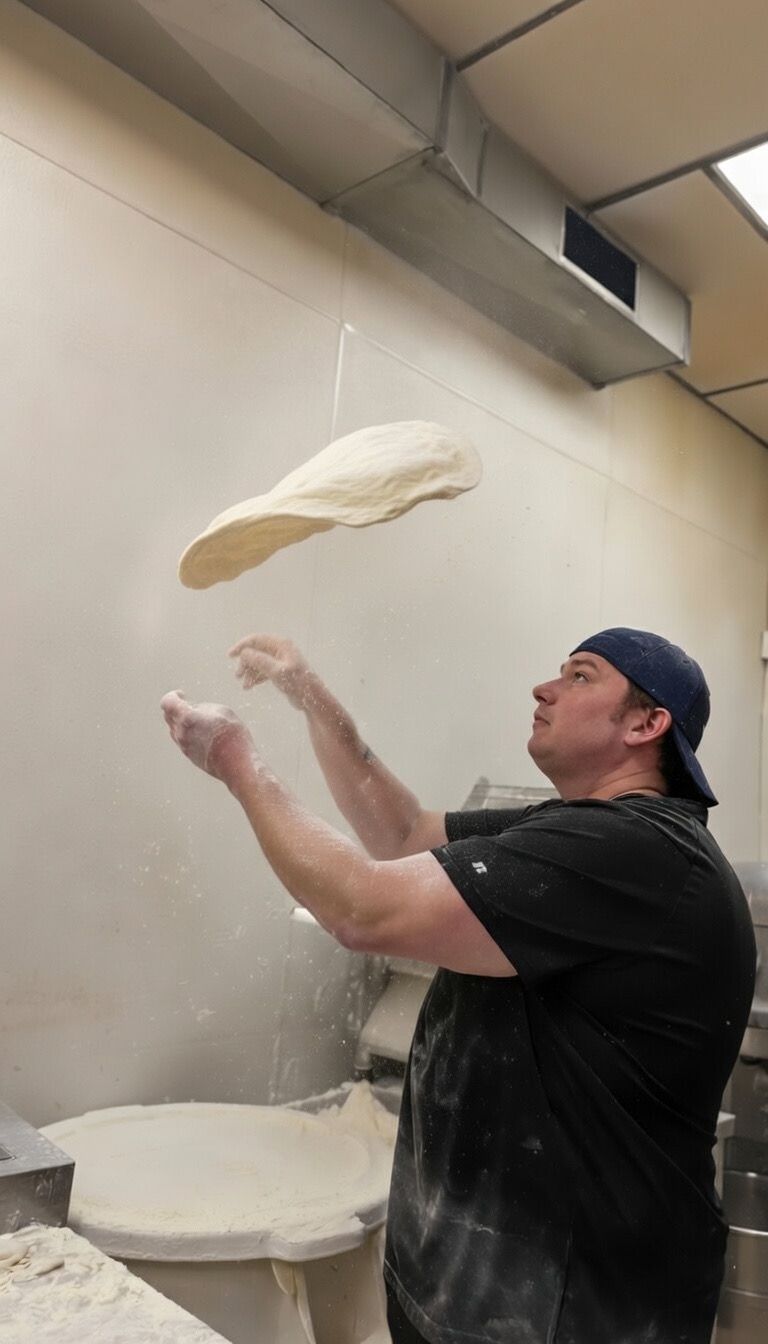 A person in a black t-shirt and baseball cap tosses pizza dough into the air in a kitchen.