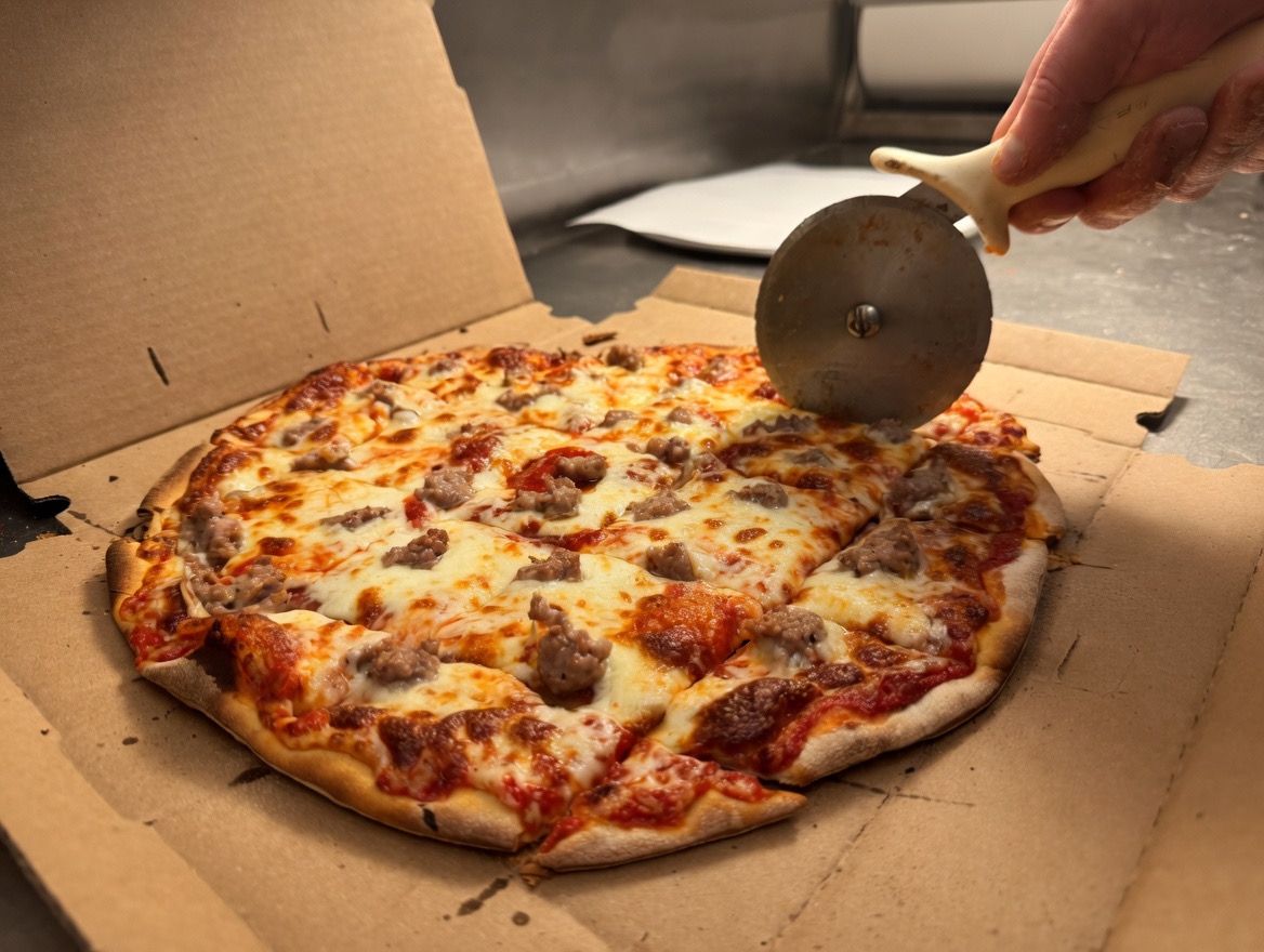 A hand uses a stainless steel rolling pizza cutter to slice a sausage pizza inside a cardboard takeout box.