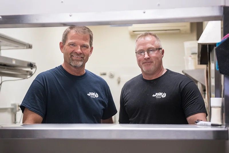 Two individuals in matching branded navy and black shirts stand side-by-side behind a stainless-steel service counter.