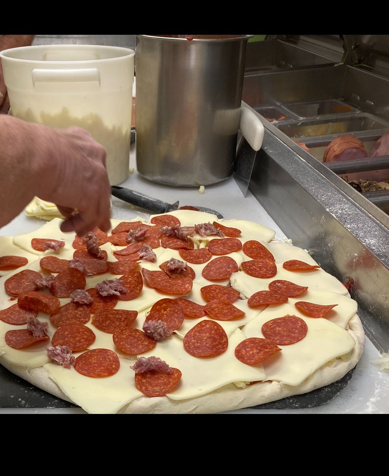 A close-up view of a person’s hand adding pepperoni and sausage toppings to a pizza covered with slices of mozzarella.