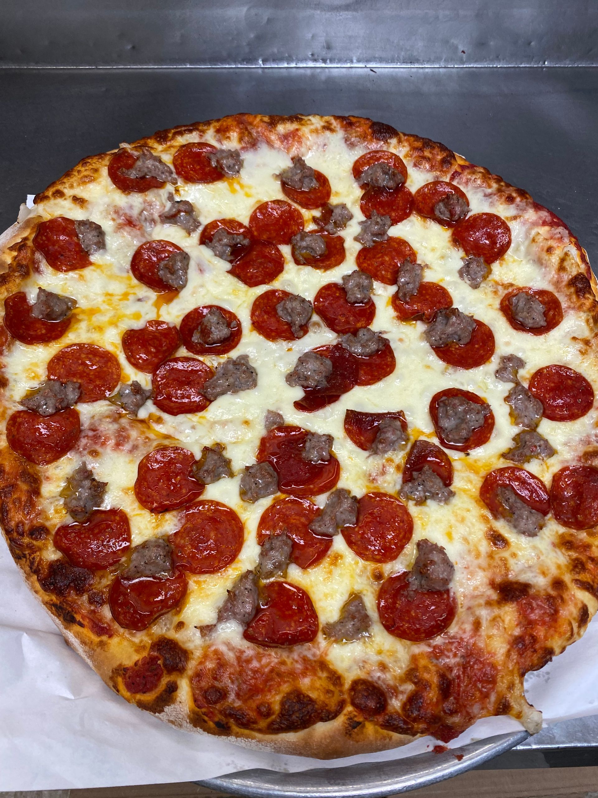A round pizza topped with pepperoni slices and sausage crumbles on a metal tray lined with white parchment paper.