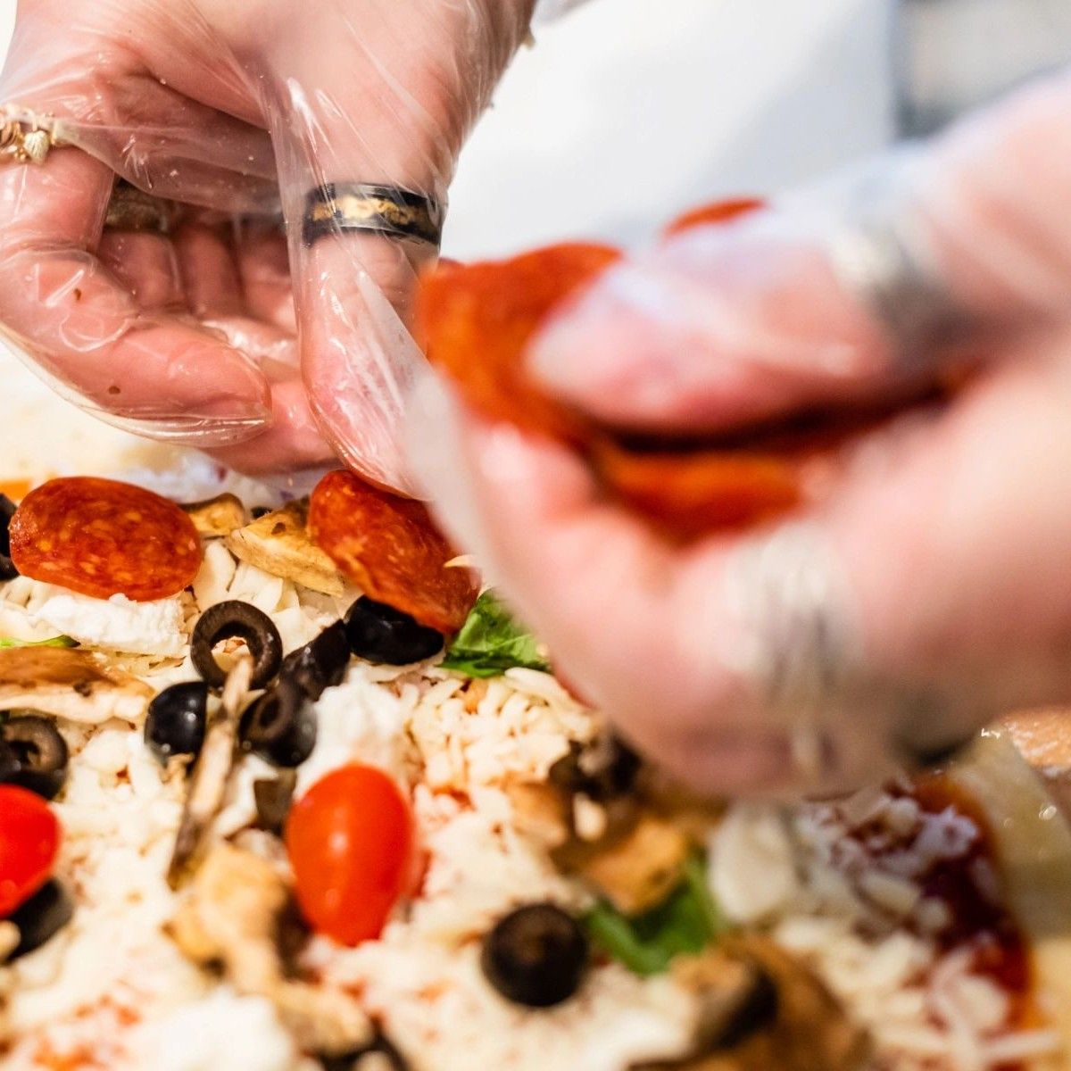 Hands in gloves placing pepperoni slices on pizza with toppings.