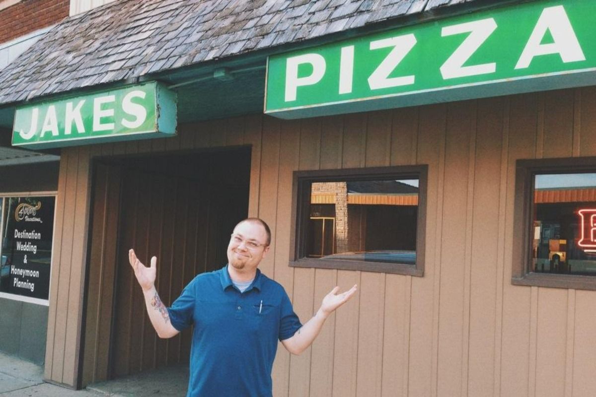 Man in blue shirt stands with arms out under the Jake's Pizza sign on a wooden building.