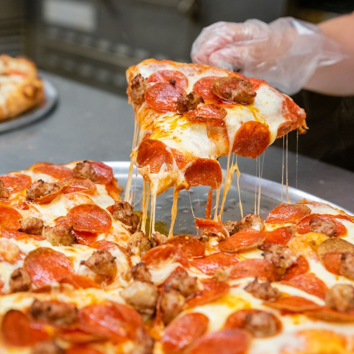 Pizza with pepperoni and sausage, cheese stretching, being held by a gloved hand.