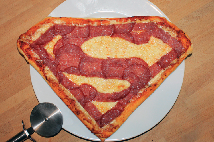 Superman logo pizza with pepperoni on a white plate, pizza cutter in view.