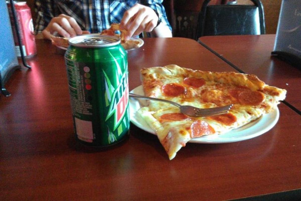 Pizza and Mountain Dew on a table. A person eats pizza in the background.