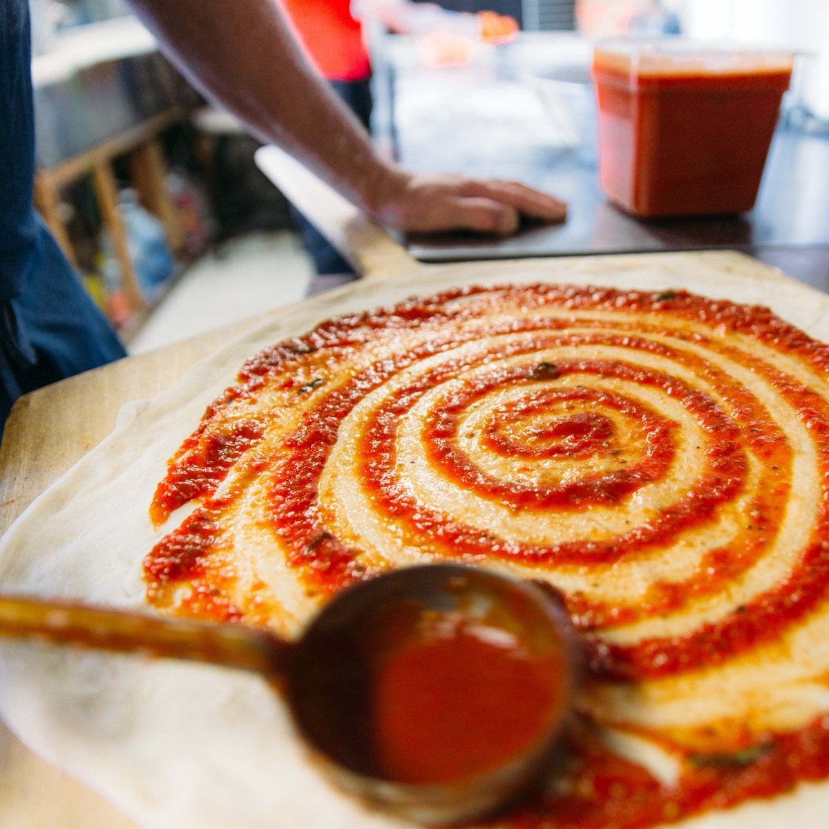 Spiral-shaped pizza with red sauce on a wooden peel, ladle of sauce in foreground, cook in background.