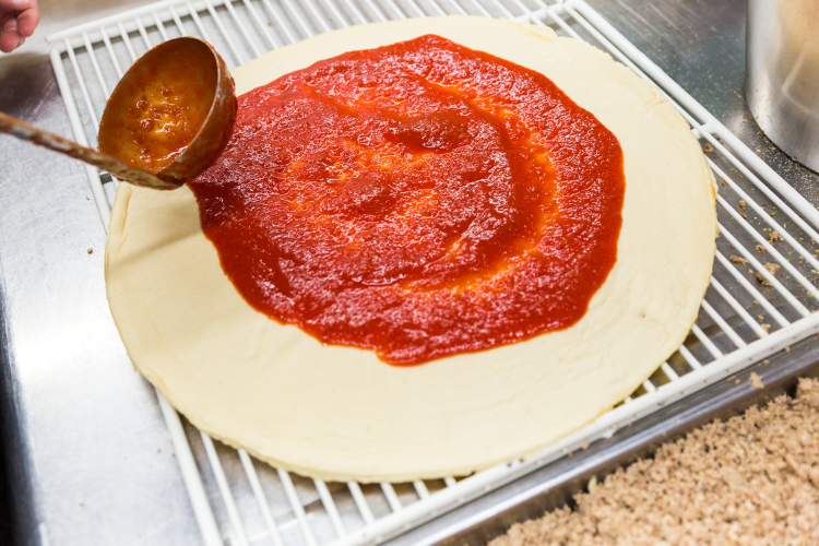 Pizza dough being sauced with a ladle on a white rack. Red sauce, cream dough.