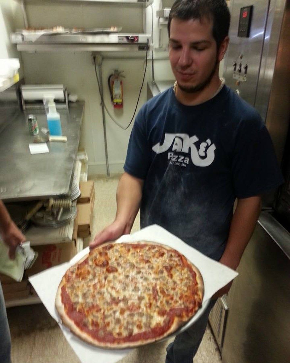 Man holding a pizza on a tray in a pizza restaurant. He wears a blue shirt with a logo.