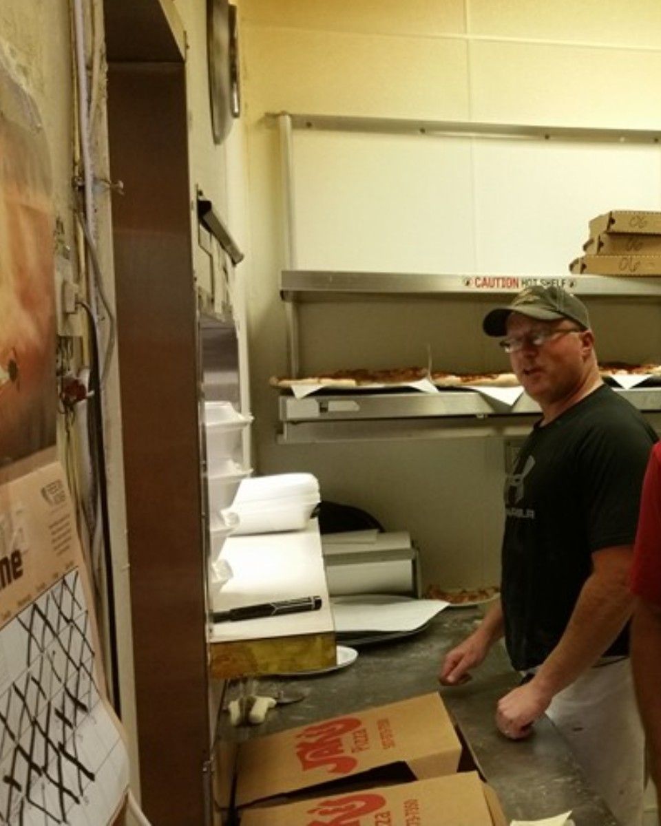 Man in a pizza kitchen, wearing a hat, standing near pizza boxes and oven.