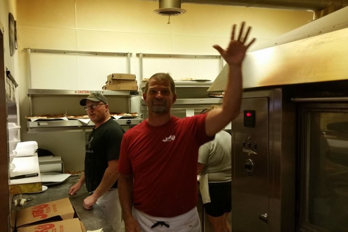 Man in red shirt waves in a kitchen, with another man and oven visible.