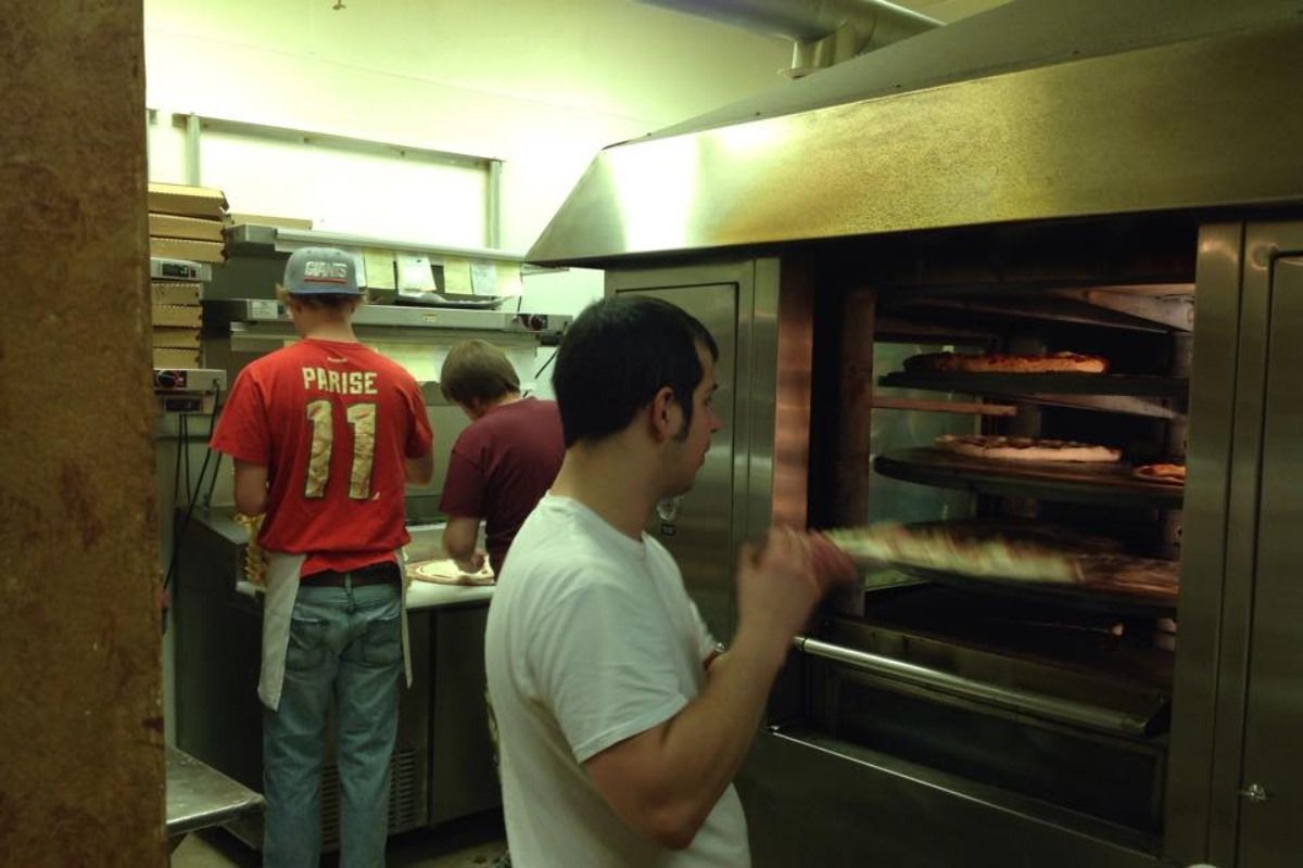 Three pizza chefs working in a kitchen, one placing a pizza in a large oven.