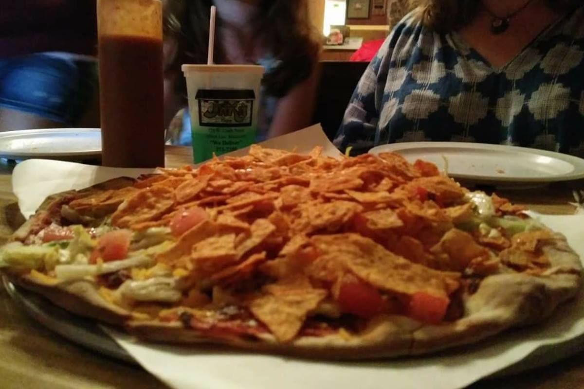 Pizza topped with tortilla chips, near drinks and two people.