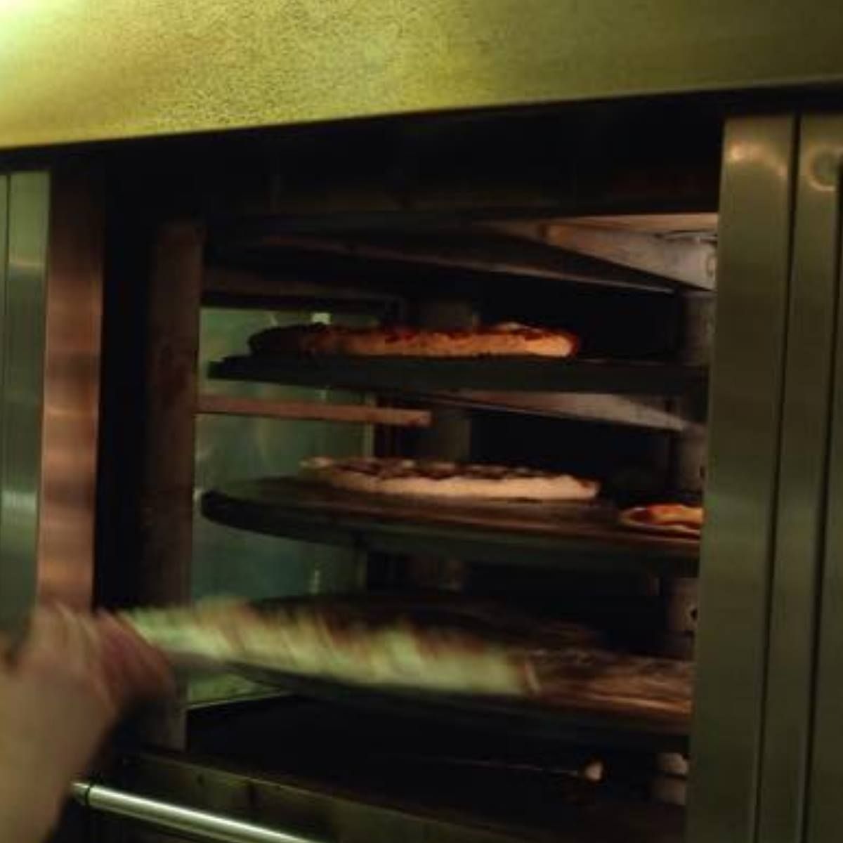 Pizzas baking on shelves inside a commercial oven, seen from the open door. A hand reaches in.